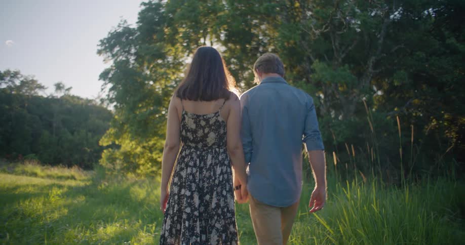 Blonde man wearing in blue shirt and brown pants hold a womans hand wearing dress in the countryside as the sun shining though trees