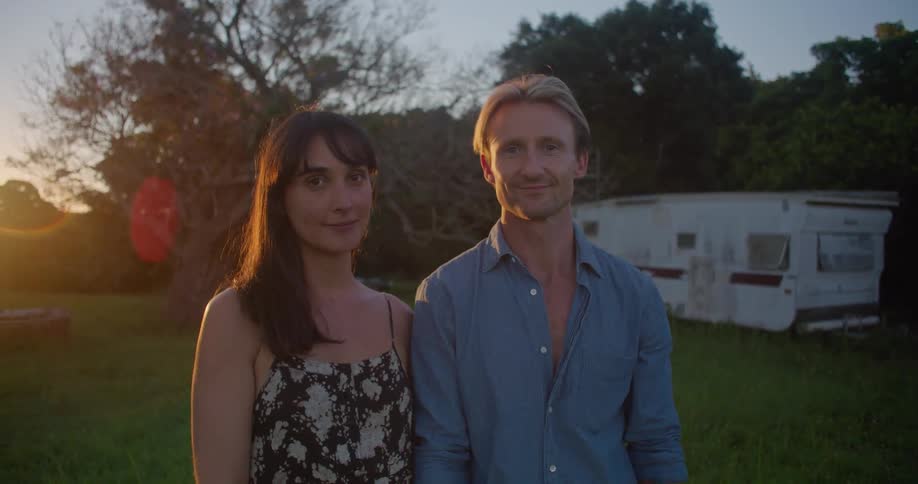 Young couple standing on a field near a trailer looking at the camera at sunset