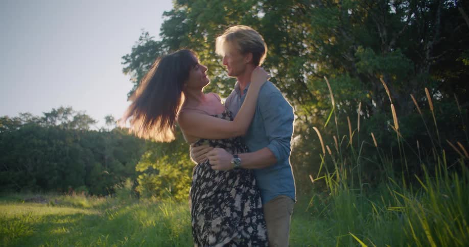 A man is carrying a woman outdoors and sets her down while they embrace and kiss in the evening