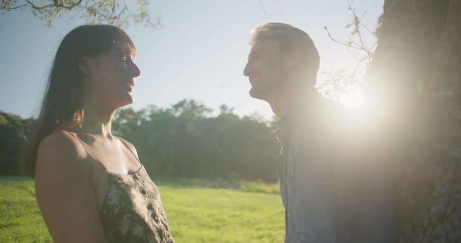 Blonde man with blue shirt leans against tree and kissing woman with wavy hair and dress holding hands