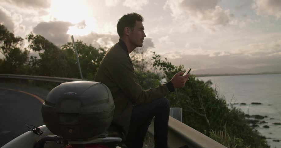 Man sits on side of road near rail overlooking ocean with surfboard texting on mobile phone