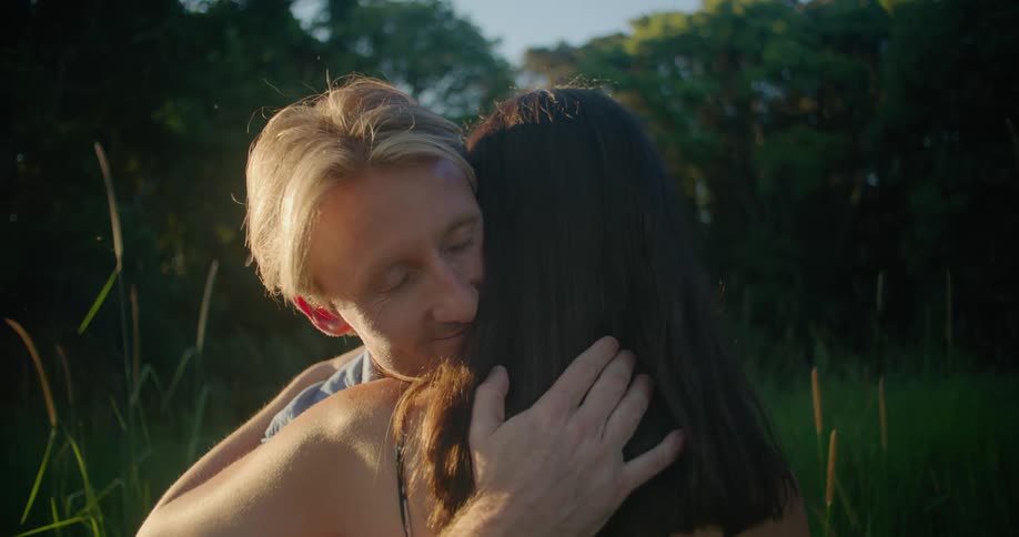 Blonde man with blue shirt cuddles woman with wavy hair and dress in the countryside