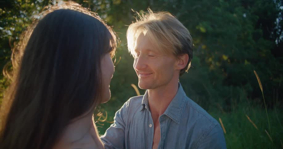 Blonde man with blue shirt and woman with brown wavy hair lovingly look into each others eyes in the countryside