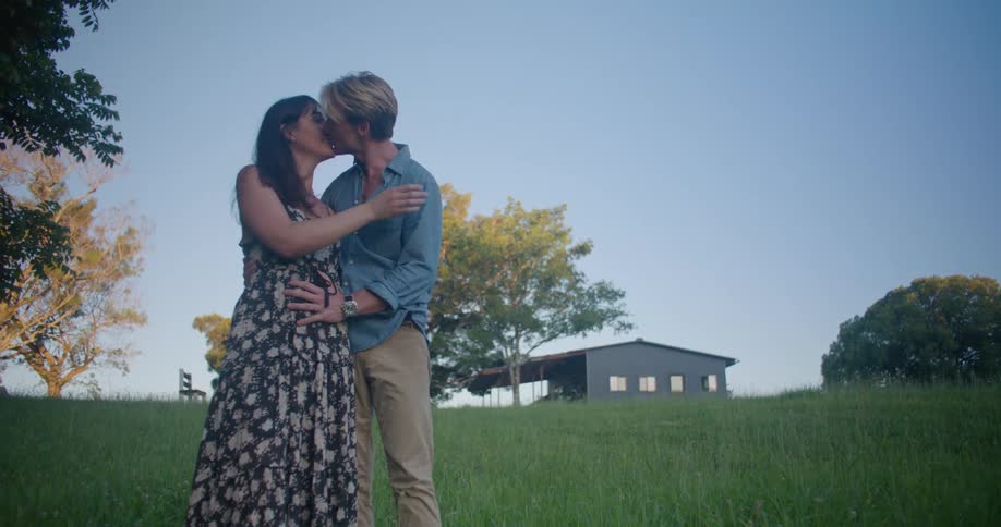 Blonde man in blue shirt and brown pants kisses woman and strokes womans belly in the countryside with country house in the background