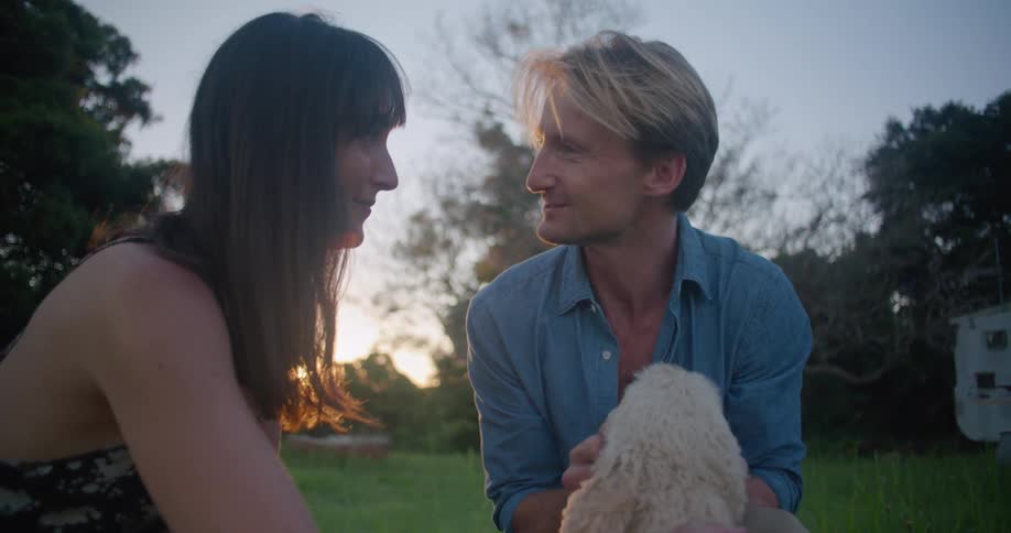 A man and woman are smiling while petting a dog outside on a lawn at sunset