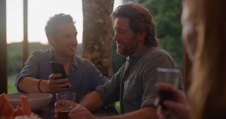Friends sit around table outdoors with drinks and watermelon laughing and talking over mobile phone