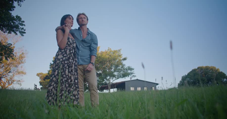 Young couple hugging and looking at the horizon while standing on a grass field at evening