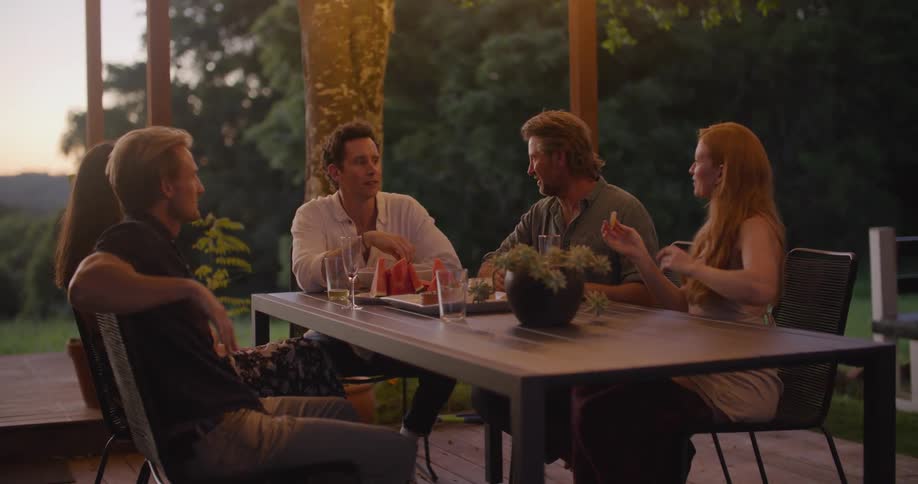 A group of friends sitting at table and talking with each other on terrace in countryside at dusk