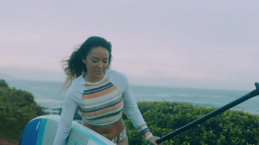 A woman carrying a surfboard along the coast places it on the ground and turns to smile at someone out of frame