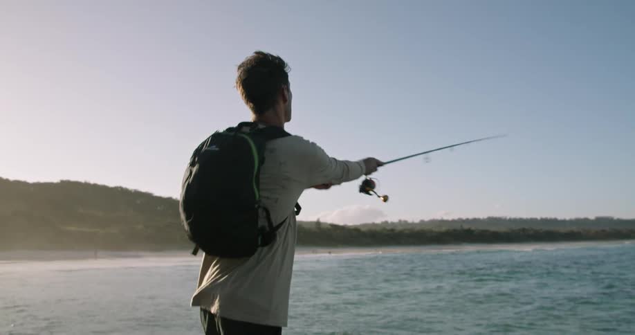 Young man fly fishing on the coast next to the ocean at sunset