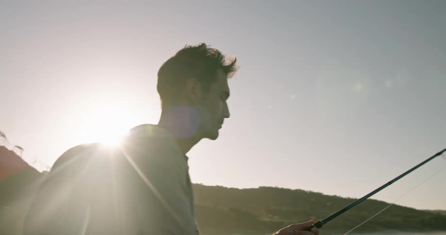 Young man fly fishing near the coast next to the mountains at sunset