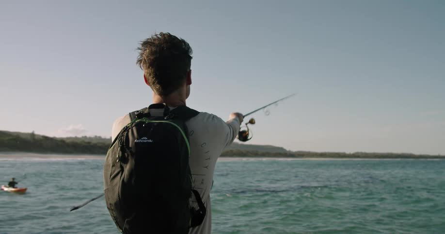 Man wearing backpack casts fishing line into ocean waters as kayaker paddles in background