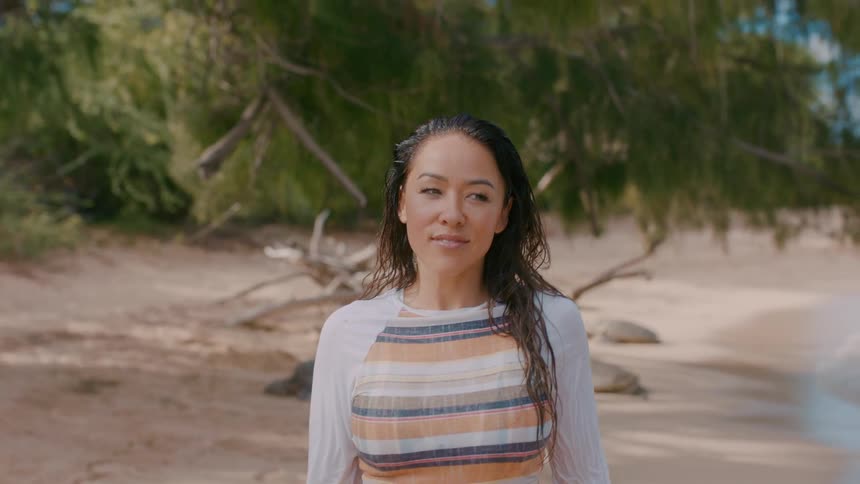 A woman with long dark hair walks across a beach with trees in the distance