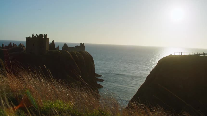 Castle ruins stand on promontory overlooking ocean across from second promontory topped with rail
