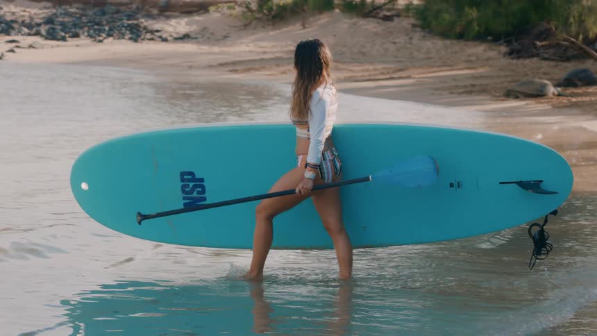 Bikini wearing woman carrying board and paddle walks into surf