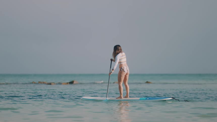 Woman stands on wake board paddling it across ocean waves