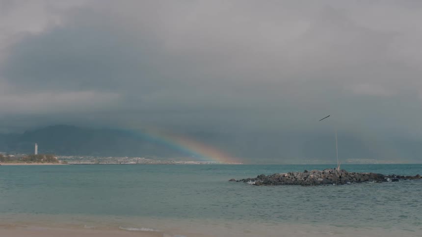 Coastal view of a small island in the water with a lighthouse in the distance and a rainbow in the sky