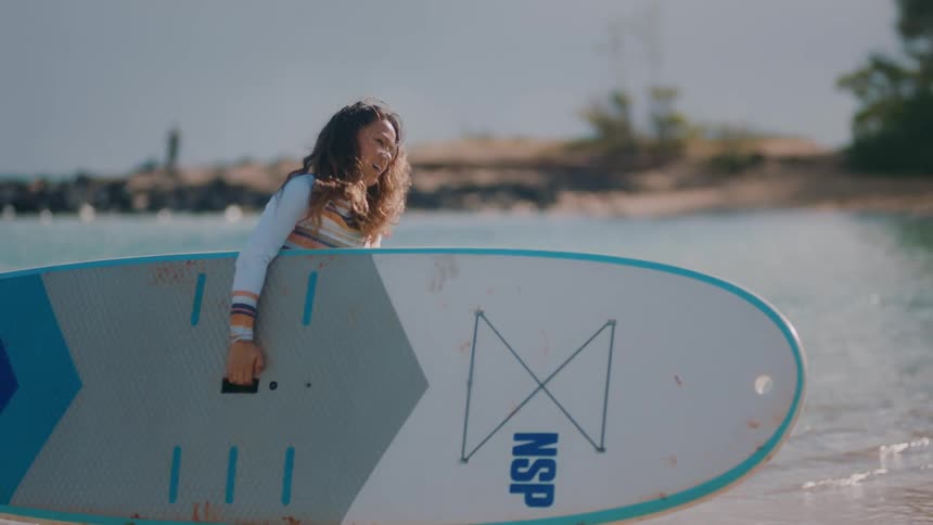 Woman carries paddle board and oar onto beach and out of water