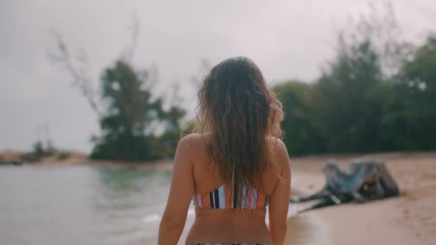 Woman wearing striped bikini walks along beach toward trees