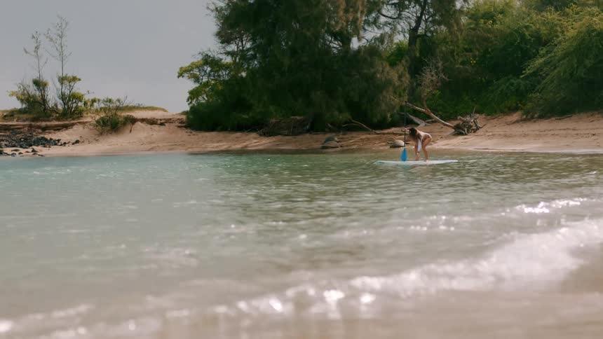 A woman kneeling atop a paddle board uses her paddle to help her stand as the board floats away from the shore