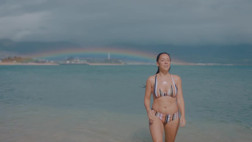 Brunette woman with bikini walks in sea and touches her wet hair with rainbow in the background