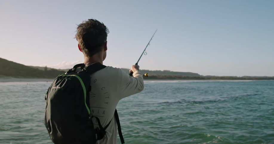 Young man fly fishing on the ocean in a coast next to the mountains at sunset