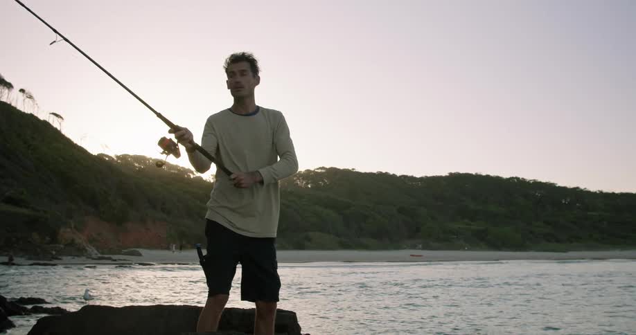 A fisherman standing on a rock on the coast casts his fishing line into the ocean in the evening
