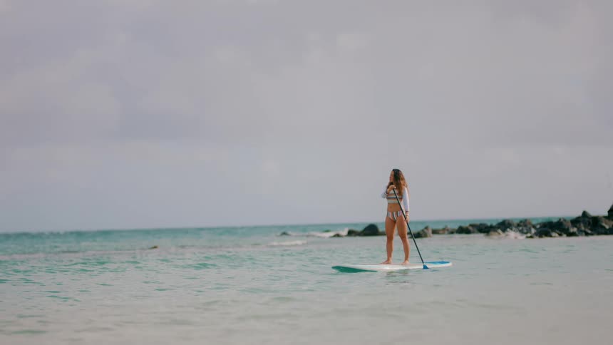 Young woman wearing sports swimwear stands on a surfboard on the ocean paddling