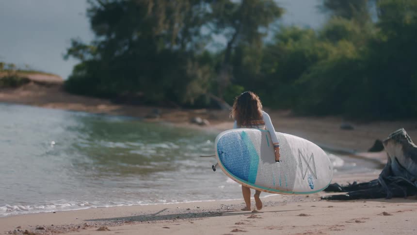 A woman carries a paddle board and paddle while walking across a beach towards the ocean