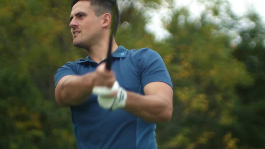 Man wearing blue shirt swings a golf club near the tree line