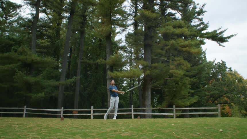Man takes shot with golf club while standing near wooden fence near woods