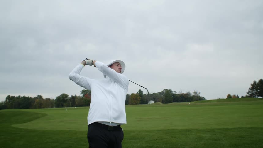 A man wearing a white hat is golfing and he swings his club hitting the ball