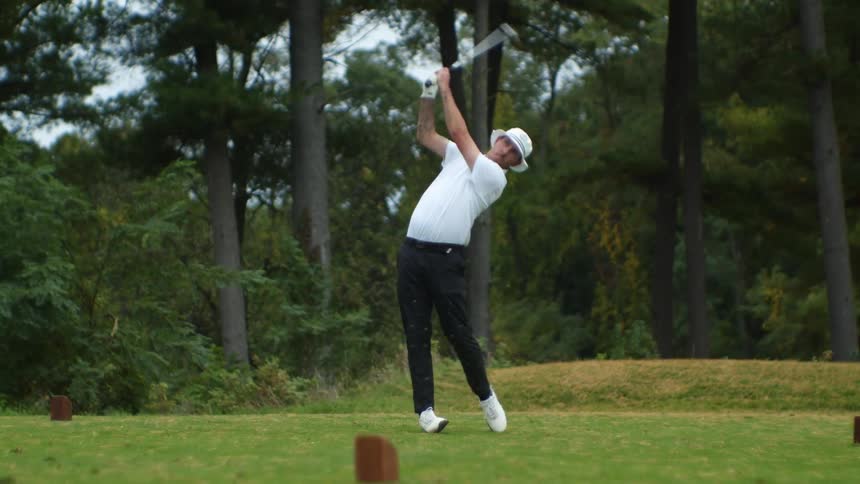 A man wearing a white hat is golfing and he swings his club hitting the ball