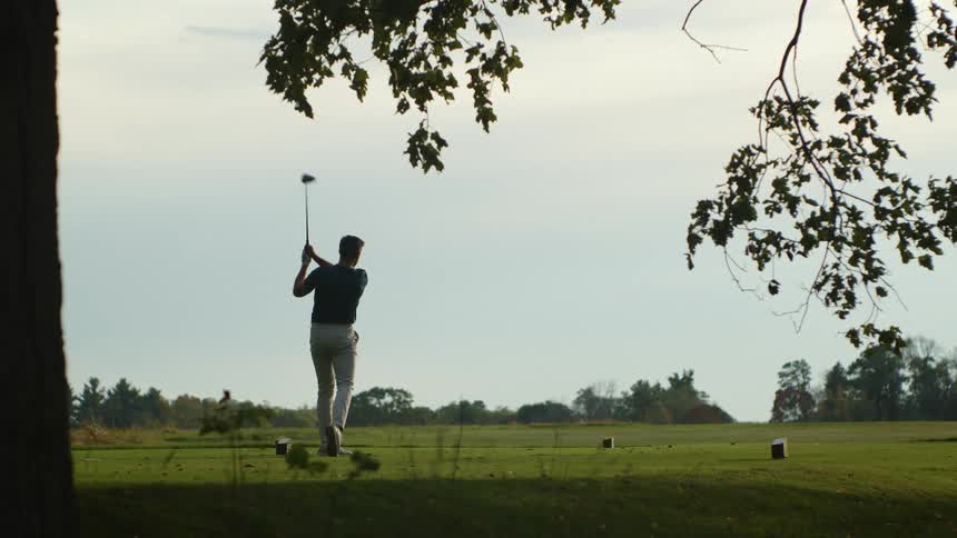 A man is on a golf course and he swings his club hitting the ball high into the sky