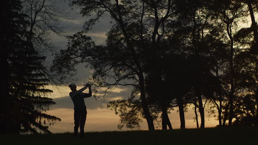 A man is hitting a golf ball with a club on a cloudy evening after sunset