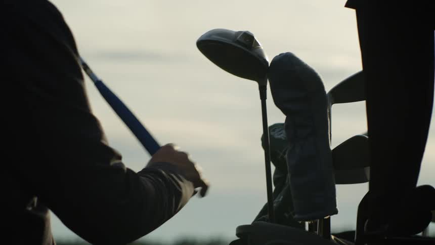 Silhouette of a man wearing a baseball hat storing golf clubs at sunset