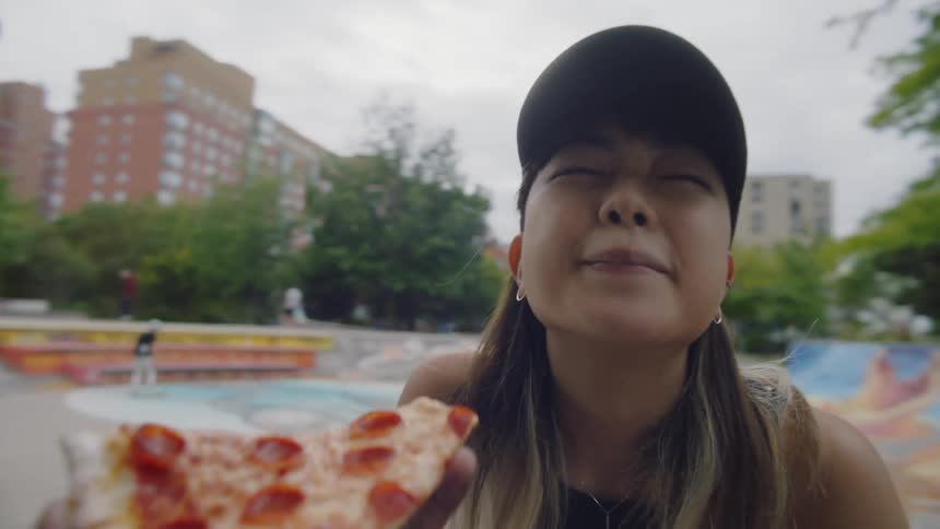 A woman smiles as she takes a bite of pepperoni pizza held by her boyfriend at the skate park