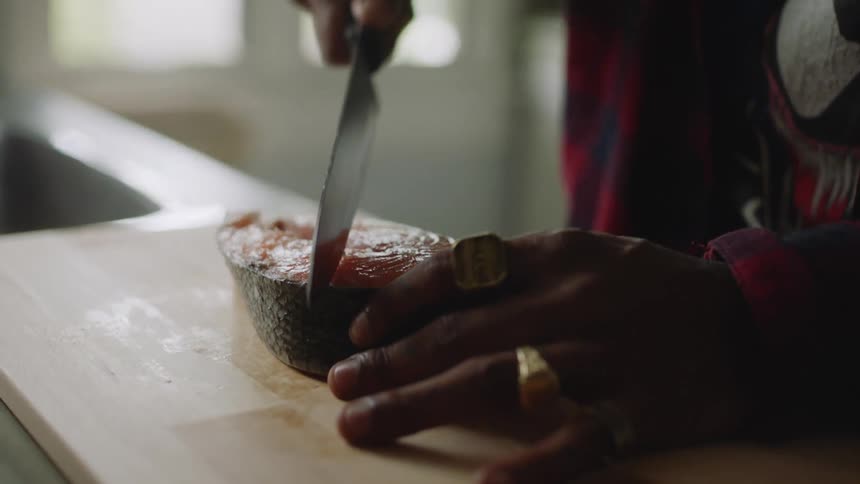 A man is standing over a piece of meat on a cutting board and puts a knife on top of the meat then stops and sets down the knife