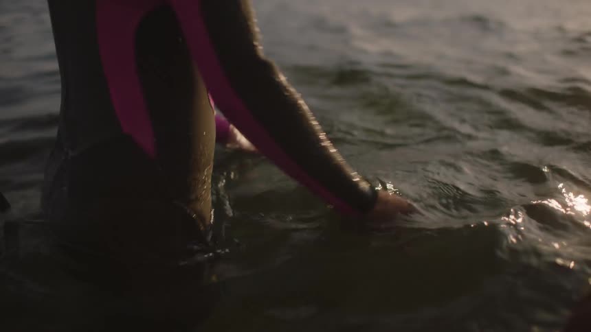 A man and a woman are wearing wetsuits and moving their hands in the water as they look at each other and smile
