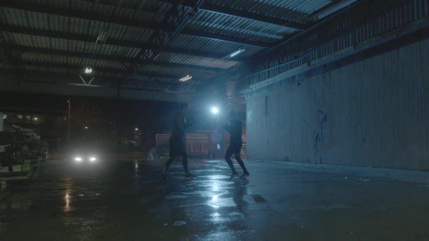 Two people are boxing in a warehouse at night with a bright light shining in the distance