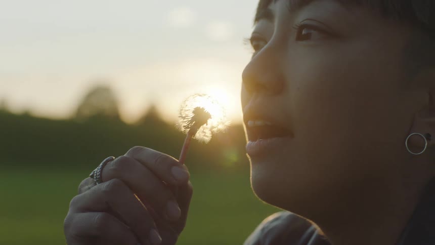A woman purses her lips and blows on a dandelion at sunset