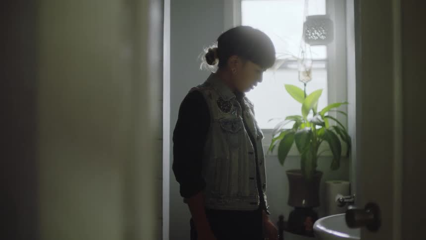 A teenage girl stands in a bathroom in a home and looks down as she flushes a toilet and then walks out of the bathroom