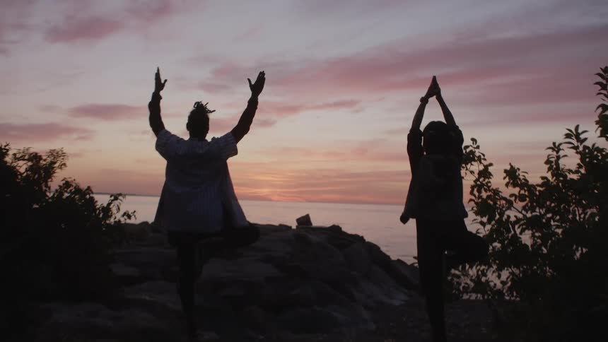 Man and woman doing yoga on the edge of cliff are silhouetted against the red sky at dusk