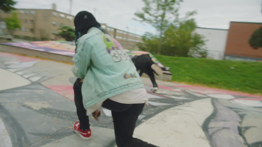 Two teenagers dance in a park as people watch and skateboard in the background