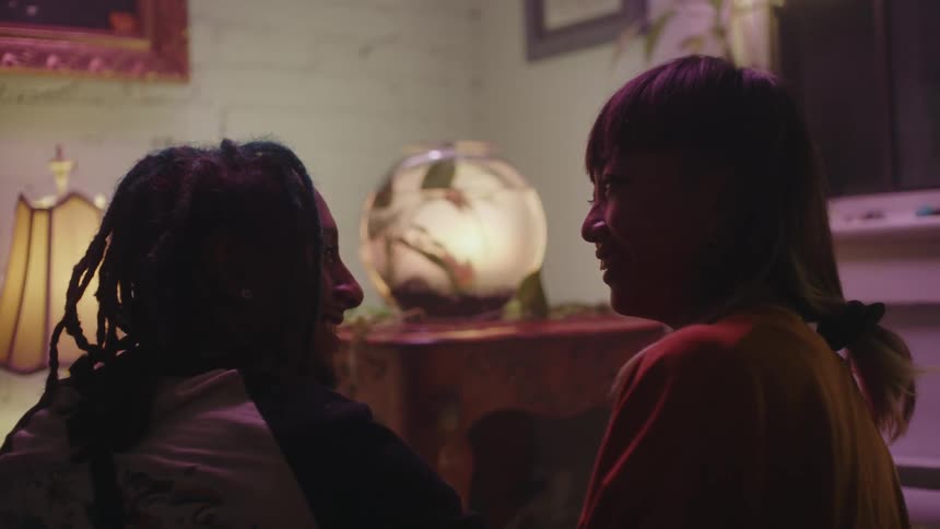 Man with braids and woman with straight hair look at glass fish bowl on table and at each others eyes in room