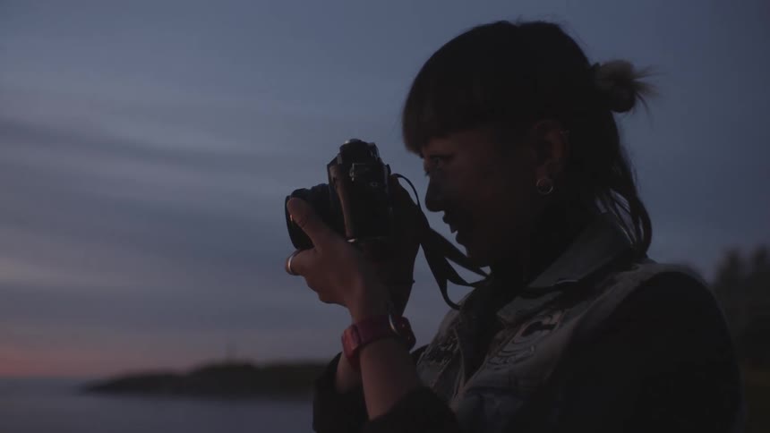A woman is standing while taking a photo of the sun setting from a beach at dusk