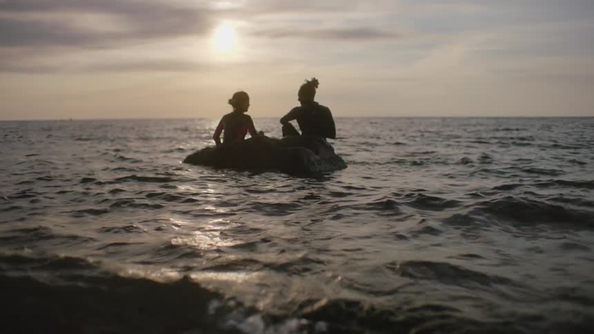 Woman and friend sit on boulder in water silhouetted by sun