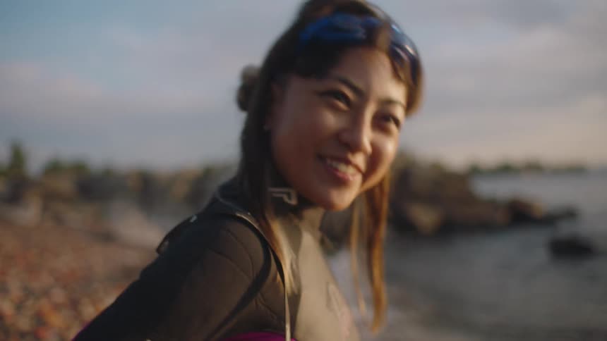 A woman wearing a wet suit and goggles on her head laughs and smiles as she has an animated conversation on shore