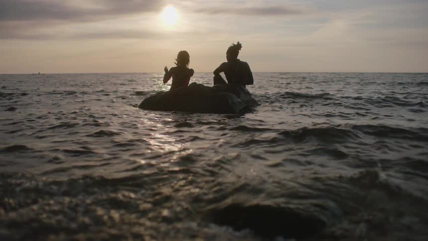 Man and woman wearing wet suits sit on large rock as waves roll past and onto shore