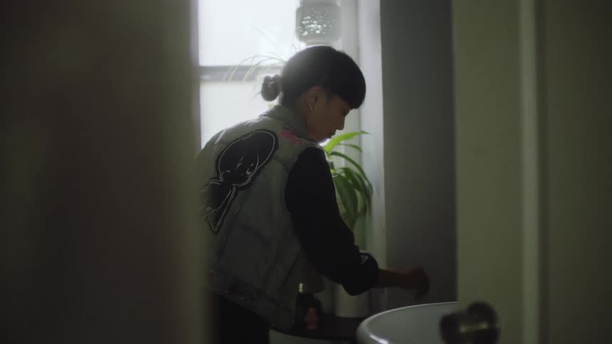 A teenage girls washes her hands in a bathroom in a home and then reaches over and looks down as she flushes a toilet